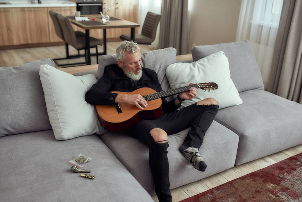 Let music flow. Bearded middle-aged man, artist writing song, playing guitar, sitting on the couch. Metal pipe for smoking marijuana, cannabis buds in a plastic bag and lighter lying on the couch - Photo, Image