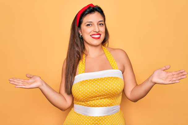 Junge hispanische Pin-up-Frau trägt Mode sexy 50er Jahre Stil über gelbem Hintergrund lächelnd zeigt beide Hände geöffnete Handflächen, präsentiert und Werbung Vergleich und Balance - Foto, Bild