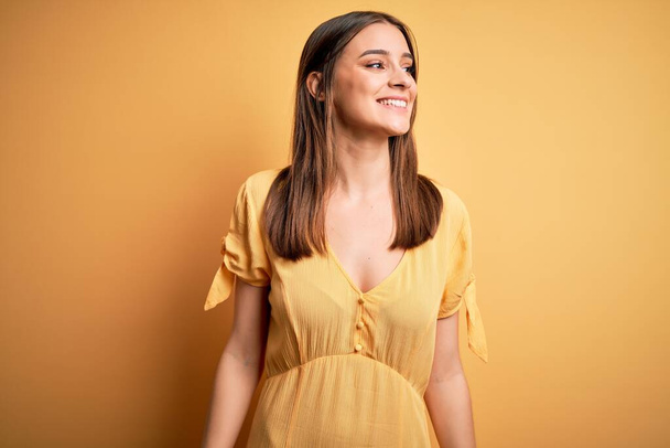 Young beautiful brunette woman wearing casual dress standing over yellow background looking away to side with smile on face, natural expression. Laughing confident. - Photo, image