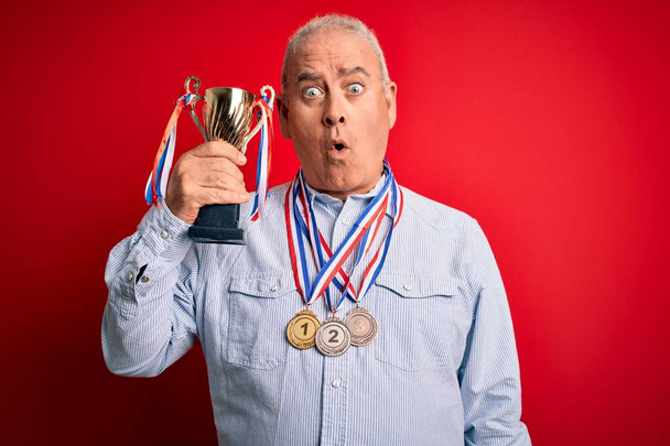 Ein erfolgreicher Mann mittleren Alters, der Medaillen mit Trophäe vor isoliertem roten Hintergrund trägt, erschrak vor Schock mit einem überraschten Gesicht, ängstlich und aufgeregt mit ängstlichem Ausdruck - Foto, Bild