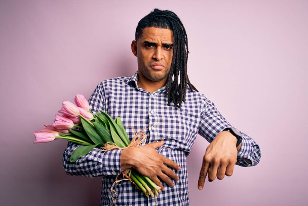Młody afrykański afro romantyczny mężczyzna z dredami trzymający bukiet różowych tulipanów wskazujący smutny i zdenerwowany, wskazujący kierunek palcami, nieszczęśliwy i przygnębiony. - Zdjęcie, obraz