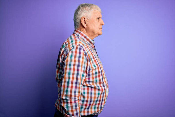 Senior hezký chraptivý muž na sobě ležérní barevné tričko přes izolované fialové pozadí při pohledu na stranu, relaxační profil póza s přirozenou tváří a sebevědomý úsměv. - Fotografie, Obrázek