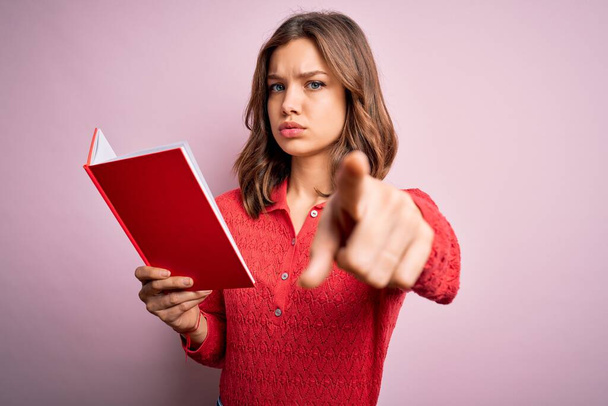 Young blonde student girl reading a book over pink  isolated background pointing with finger to the camera and to you, hand sign, positive and confident gesture from the front - Photo, Image