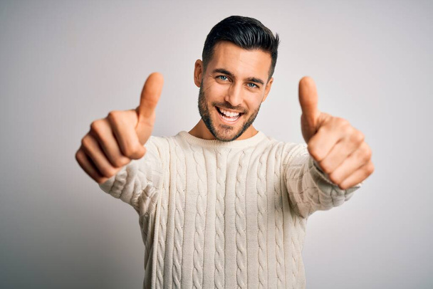 Young handsome man wearing casual sweater standing over isolated white background approving doing positive gesture with hand, thumbs up smiling and happy for success. Winner gesture. - Foto, immagini