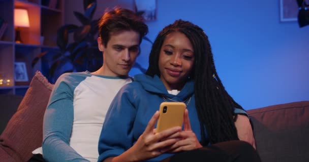 Front view of young happy multiethnic couple looking at smartphone screen while sitting on sofa. Millennial boyfriend and girlfriend browsing internet while spending time together at home. - Footage, Video