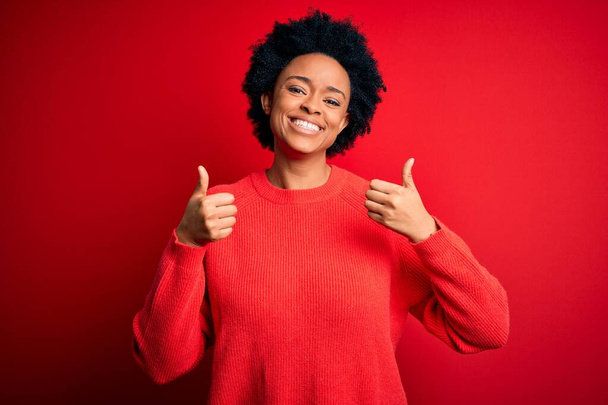 Jonge mooie Afro-Amerikaanse afro vrouw met krullend haar dragen casual trui succes teken doet positieve gebaar met de hand, duimen omhoog glimlachend en gelukkig. Vrolijke uitdrukking en winnaar gebaar. - Foto, afbeelding