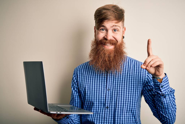 Kızıl saçlı, sakallı bir İrlandalı iş adamı bilgisayarlı dizüstü bilgisayarını izole edilmiş arka planda kullanıyor. Bir fikir ya da soru işareti ile şaşkın bir şekilde mutlu yüzü işaret ediyor. - Fotoğraf, Görsel