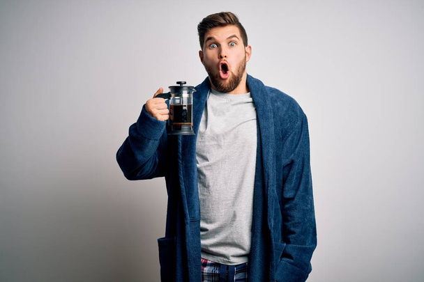 Junger blonder Mann mit Bart und blauen Augen, der Pyjama trägt und Kaffee mithilfe der Kaffeemaschine kocht, erschrocken mit einem Überraschungsgesicht, ängstlich und aufgeregt mit Angstausdruck - Foto, Bild