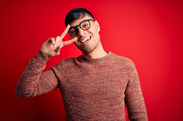 Giovane uomo ispanico bello indossa occhiali nerd su sfondo rosso Fare il simbolo della pace con le dita sul viso, sorridente allegro mostrando la vittoria
 - Foto, immagini