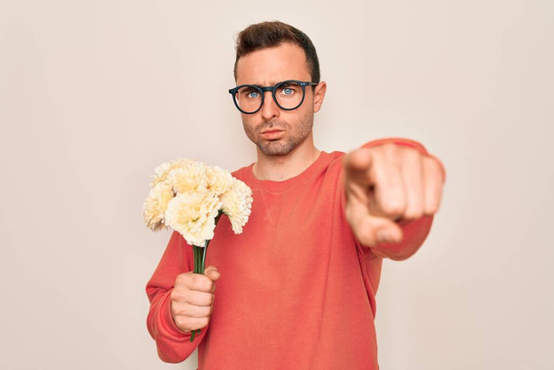 Młody przystojny mężczyzna z niebieskimi oczami trzymający bukiet kwiatów na białym tle wskazując palcem na aparat i na ciebie, znak dłoni, pozytywny i pewny gest z przodu - Zdjęcie, obraz