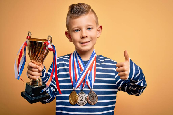 Młody mały biały dzieciak noszący medale zwycięzcy i zwycięstwo trofeum nagrody nad żółtym tle szczęśliwy z dużym uśmiechem robi znak ok, kciuk w górę palcami, doskonały znak - Zdjęcie, obraz