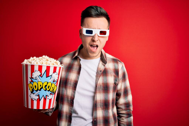 Młody przystojny biały mężczyzna w okularach 3D i jedzący popcorn w szokującej twarzy, wyglądający sceptycznie i sarkastycznie, zaskoczony otwartymi ustami - Zdjęcie, obraz