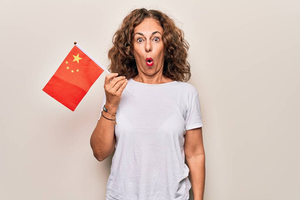 Middle age beautiful patriotic woman holding chinese flag over isolated white background scared and amazed with open mouth for surprise, disbelief face - Фото, зображення