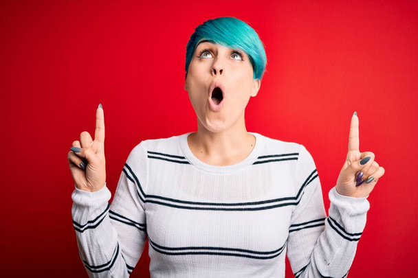 Young beautiful woman with blue fashion hair wearing striped sweater over red background amazed and surprised looking up and pointing with fingers and raised arms. - Photo, Image