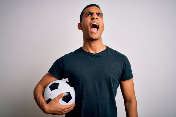 Pohledný africký Američan hrající fotbal, držící fotbalový míček nad bílým pozadím, vzteklý a šílený, frustrovaný a zuřivý, křičící vzteky. Vztek a agresivní koncepce. - Fotografie, Obrázek