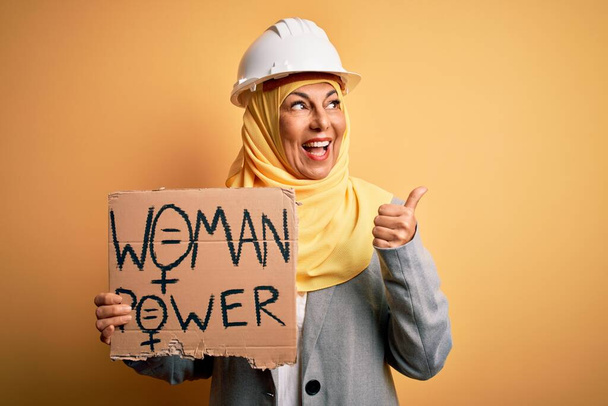 Жінка-архітектор середнього віку в мусульманському хіджабі тримає прапор з жіночим повідомленням, вказуючи і показуючи з великим пальцем в сторону з щасливим обличчям посміхаючись
 - Фото, зображення
