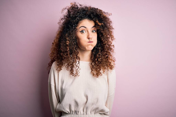Beautiful woman with curly hair and piercing wearing casual sweater over pink background puffing cheeks with funny face. Mouth inflated with air, crazy expression. - Photo, Image