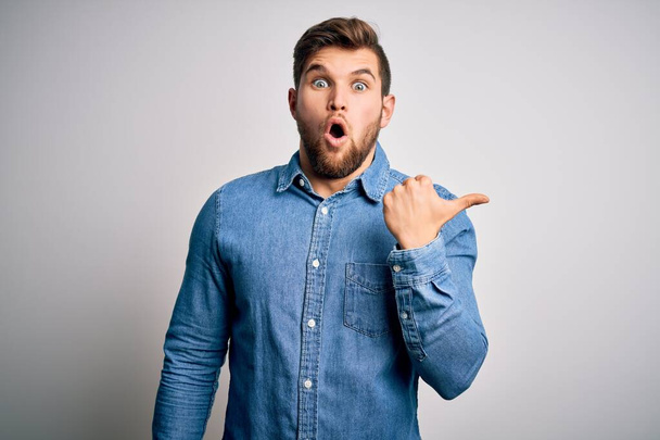Young handsome blond man with beard and blue eyes wearing casual denim shirt Surprised pointing with hand finger to the side, open mouth amazed expression. - Foto, immagini