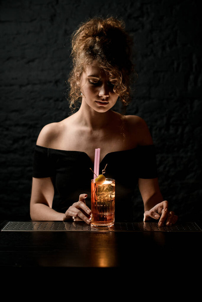 Young attractive female bartender is looking at glass with ready-made cocktail on bar. - 写真・画像