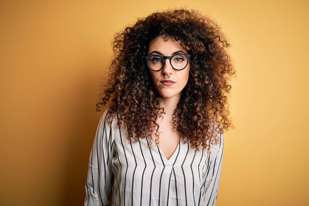 Young beautiful woman with curly hair and piercing wearing striped shirt and glasses Relaxed with serious expression on face. Simple and natural looking at the camera. - Foto, immagini