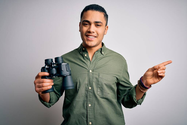 Молодий бразильський чоловік дивиться через бінокль над ізольованим білим тлом дуже щасливий, вказуючи рукою і пальцем на бік
 - Фото, зображення