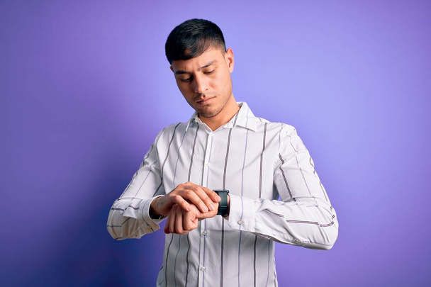 Młody przystojny Latynos w eleganckiej koszuli biznesowej stojącej na fioletowym tle Sprawdzanie czasu na zegarku, zrelaksowany i pewny siebie - Zdjęcie, obraz