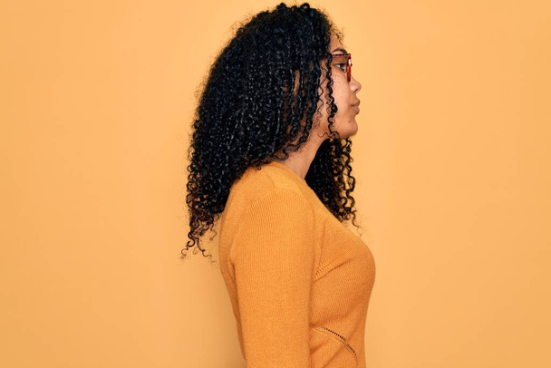 Mujer afroamericana joven con suéter casual y gafas sobre fondo amarillo mirando a un lado, pose de perfil relajante con cara natural con sonrisa confiada
. - Foto, imagen