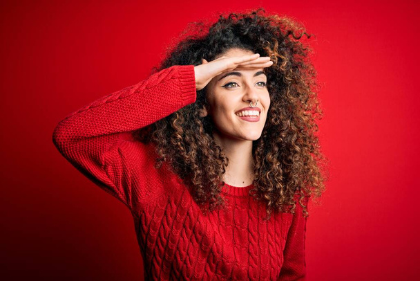 Young beautiful woman with curly hair and piercing wearing casual red sweater very happy and smiling looking far away with hand over head. Searching concept. - Photo, Image