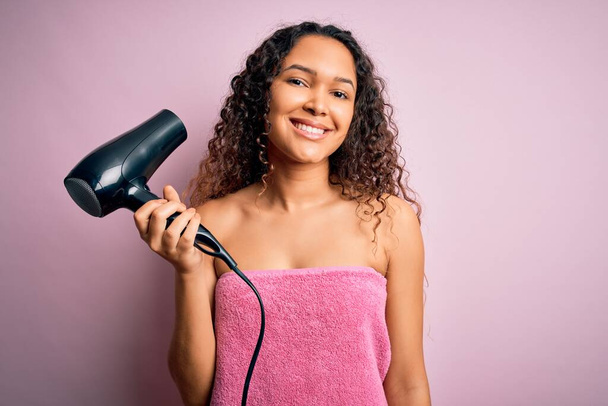 Όμορφη γυναίκα με σγουρά μαλλιά φορώντας πετσέτα μπάνιου μετά το μπάνιο χρησιμοποιώντας στεγνωτήρα μαλλιών με χαρούμενο πρόσωπο στέκεται και χαμογελά με αυτοπεποίθηση χαμόγελο που δείχνει τα δόντια - Φωτογραφία, εικόνα