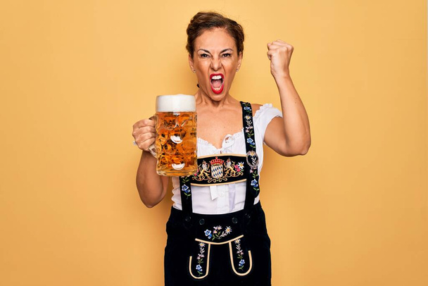 Mujer morena de mediana edad con vestido de oktoberfest tradicional alemán bebiendo tarro de cerveza molesto y frustrado gritando con ira, loco y gritando con la mano levantada, concepto de ira
 - Foto, imagen