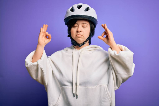 Giovane donna ciclista sindrome di Down indossando casco bici di sicurezza su sfondo viola rilassarsi e sorridere con gli occhi chiusi facendo gesto di meditazione con le dita. Concetto yoga
. - Foto, immagini