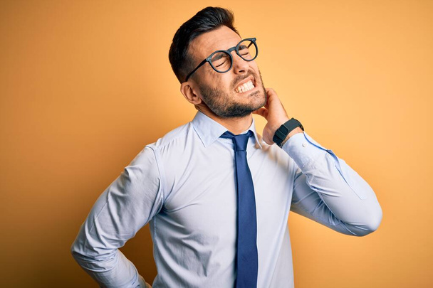 Νεαρός όμορφος επιχειρηματίας με γραβάτα και γυαλιά πάνω από κίτρινο φόντο Πόνος στον αυχένα, αγγίζοντας τον λαιμό με το χέρι, μυϊκός πόνος - Φωτογραφία, εικόνα