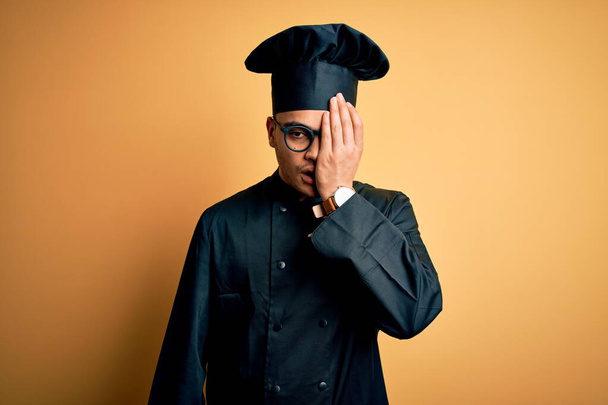 Young brazilian chef man wearing cooker uniform and hat over isolated yellow background Yawning tired covering half face, eye and mouth with hand. Face hurts in pain. - Photo, Image