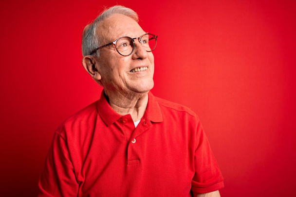 Hombre mayor de pelo gris con gafas y camiseta casual sobre fondo rojo mirando hacia un lado con sonrisa en la cara, expresión natural. Riendo confiado
. - Foto, imagen