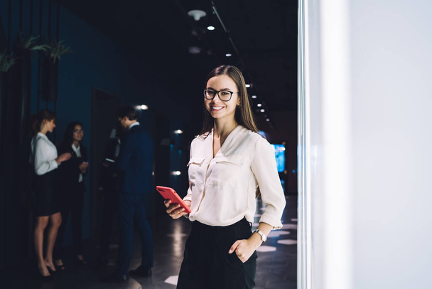 Low angle of joyful adult woman in glasses and elegant clothes smiling at camera while standing with hand in pocket and using smartphone in dark corridor of modern office against blurred pensive colleagues talking to each other - Valokuva, kuva