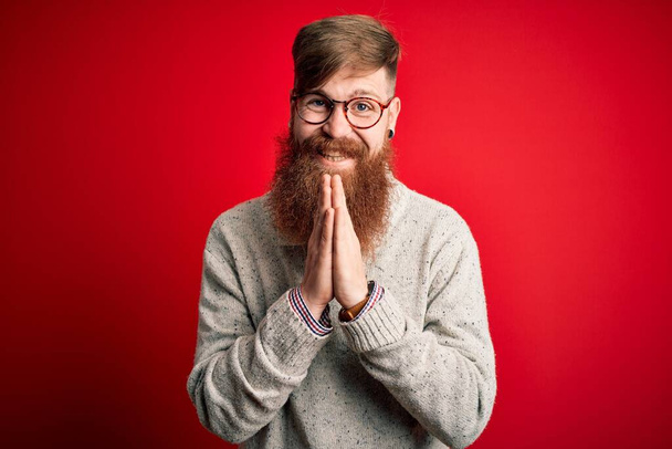 Schöner irischer Rotschopf mit Bart, lässigem Pullover und Brille vor rotem Hintergrund, der mit den Händen um Vergebung fleht und selbstbewusst lächelt. - Foto, Bild
