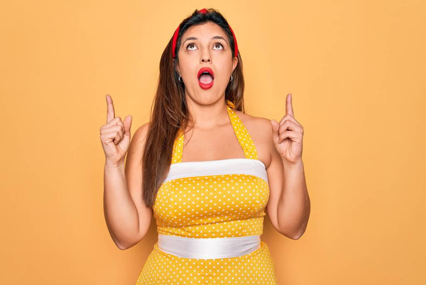 Young hispanic pin up woman wearing fashion sexy 50s style over yellow background amazed and surprised looking up and pointing with fingers and raised arms. - Foto, Imagen