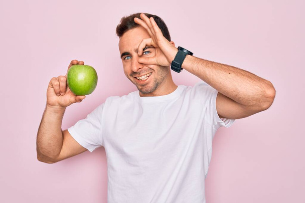 Young handsome man with blue eyes eating healthy green apple over isolated pink background with happy face smiling doing ok sign with hand on eye looking through fingers - Photo, Image