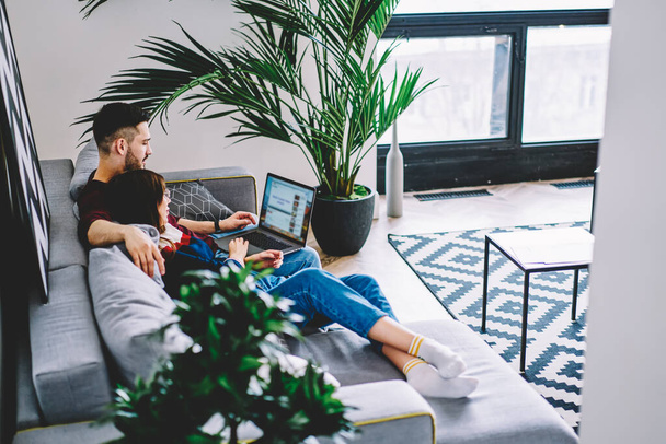 Caucasian couple in love enjoying free time on cozy sofa and watching movie on modern laptop using wireless internet connection in stylish apartment, young wife and husband resting at home interior - Fotografie, Obrázek