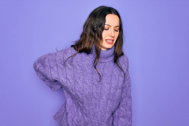 Young beautiful woman wearing casual turtleneck sweater standing over purple background Suffering of backache, touching back with hand, muscular pain - Foto, imagen