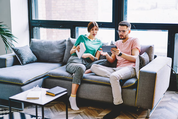 Caucasian marriage spending time together in flat with modern interior discussing information from website on touch pad, young girlfriend and boyfriend resting on comfortable couch chatting via tablet - Photo, Image