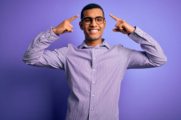 Komea afrikkalainen amerikkalainen mies yllään raidallinen paita ja lasit violetti tausta hymyillen pään molemmin käsin sormella, hyvä idea tai ajatus, hyvä muisti
 - Valokuva, kuva