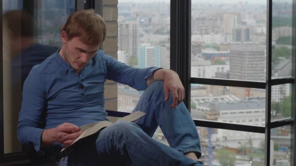 Young caucasian man sitting on windowsill, reads a book, laughs and turns the page. Activity, hobby during quarantine of coronavirus pandemic COVID-19. Self isolation, stay home concept. 4K 60fps slow motion - Filmmaterial, Video