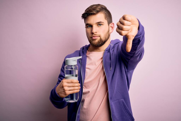 Young blond sporty man with beard and blue eyes doing sport holding bottle of water with angry face, negative sign showing dislike with thumbs down, rejection concept - Photo, image