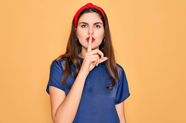 Jonge mooie brunette vrouw draagt rode lippen over gele achtergrond vragen om stil te zijn met de vinger op de lippen. Stilte en geheim concept. - Foto, afbeelding