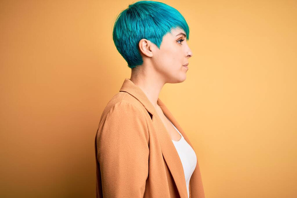 Young beautiful woman with blue fashion hair wearing casual jacket over yellow background looking to side, relax profile pose with natural face with confident smile. - Foto, Imagem