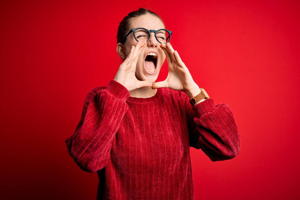 Young beautiful redhead woman wearing casual sweater over isolated red background Shouting angry out loud with hands over mouth - Photo, Image