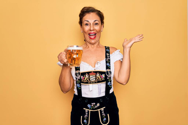 Keski-ikä brunette nainen yllään saksalainen perinteinen oktoberfest mekko juominen purkki olutta erittäin onnellinen ja innoissaan, voittaja ilme juhlii voittoa huutaen iso hymy ja nosti kädet - Valokuva, kuva