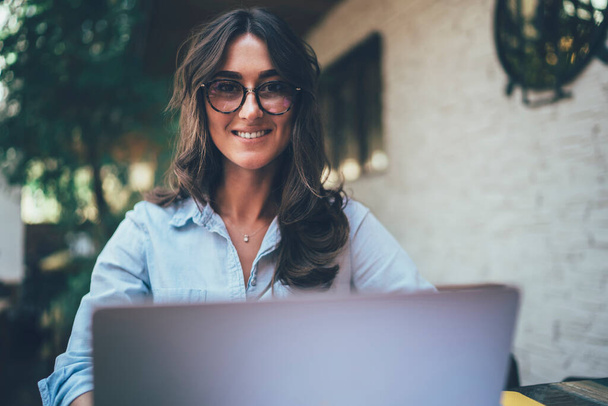 Low angle of happy adult lady in glasses and casual clothes smiling and looking at camera while sitting at table and using laptop on cafe terrace in daylight - Photo, image