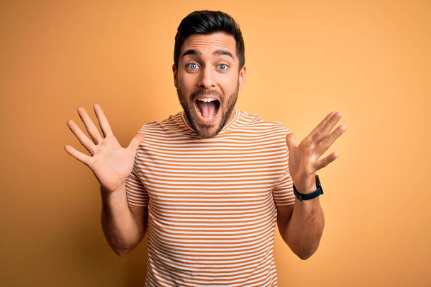 Young handsome man with beard wearing casual striped t-shirt over yellow background celebrating crazy and amazed for success with arms raised and open eyes screaming excited. Winner concept - Fotografie, Obrázek